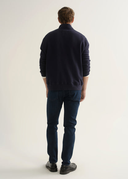 Person wearing a navy long-sleeve pullover with a mock collar, dark indigo slim tapered jeans, and black leather shoes, back full body view on a light grey background.