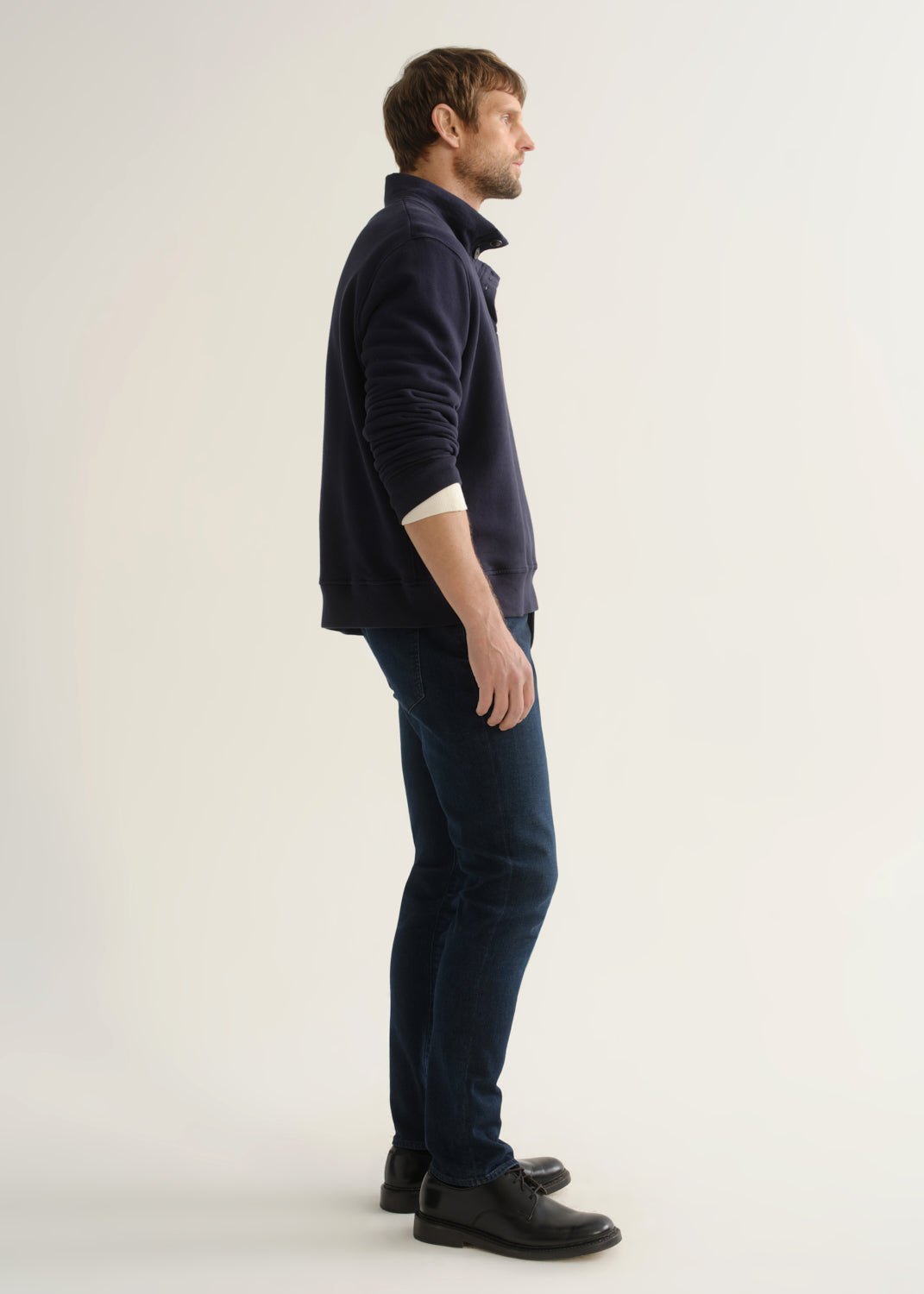Person wearing a navy long-sleeve pullover with a mock collar and button placket layered over a white tee, dark indigo slim tapered jeans, and black leather shoes, side full body view on a light grey background.