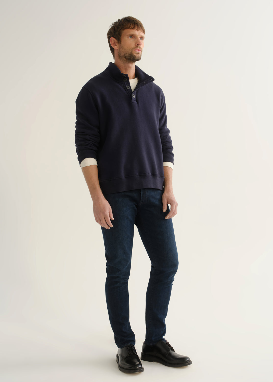 Person wearing a navy long-sleeve pullover with a mock collar and button placket layered over a white tee, dark indigo slim tapered jeans, and black leather shoes, front full body view on a light grey background.