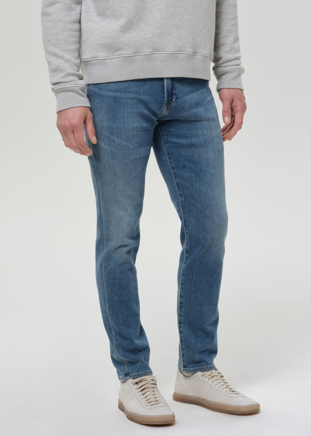 Man wearing medium blue straight leg jeans with a light grey crewneck sweatshirt on a grey background.