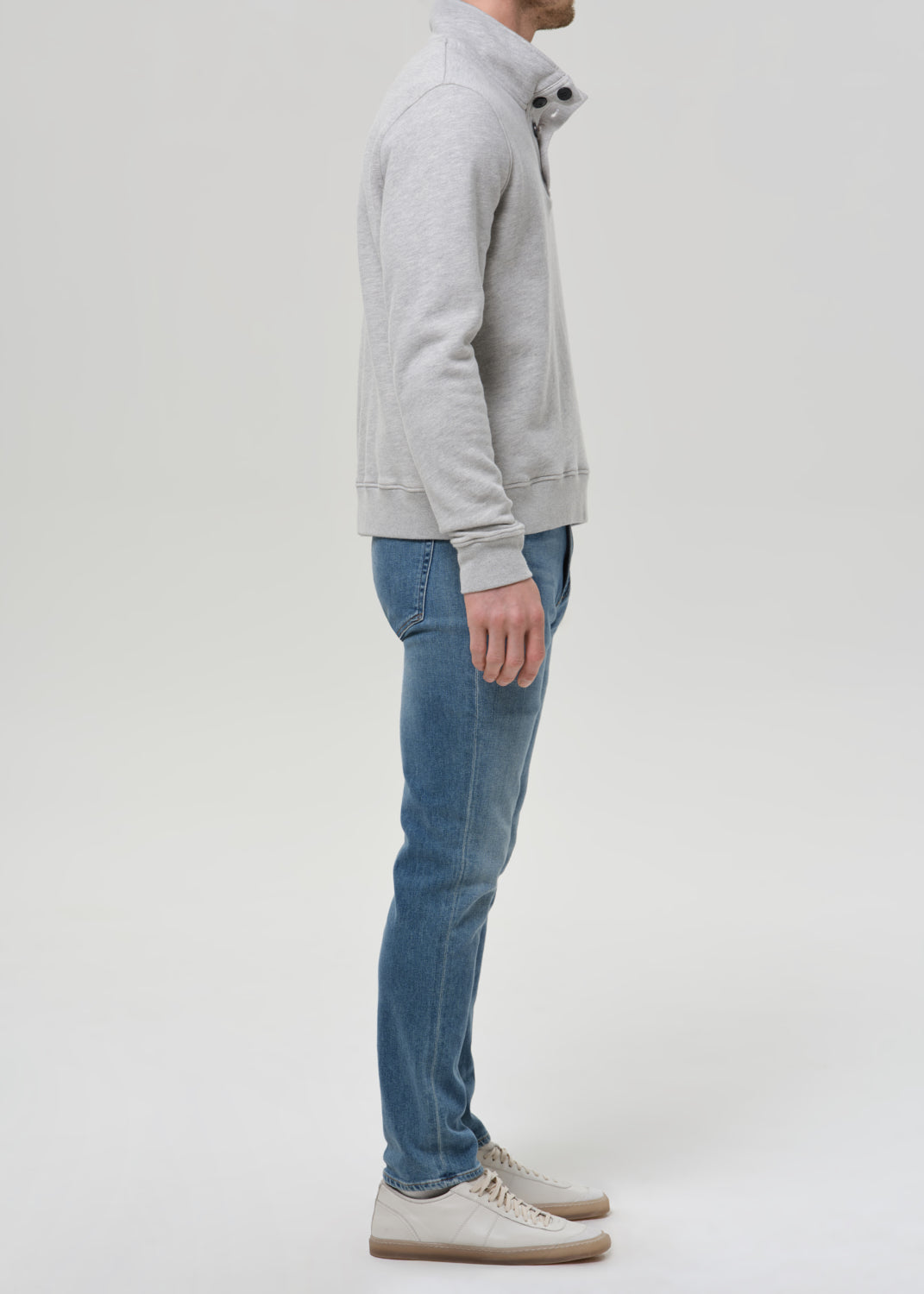 Man wearing medium blue straight leg jeans with a light grey crewneck sweatshirt viewed from the side on a grey background.