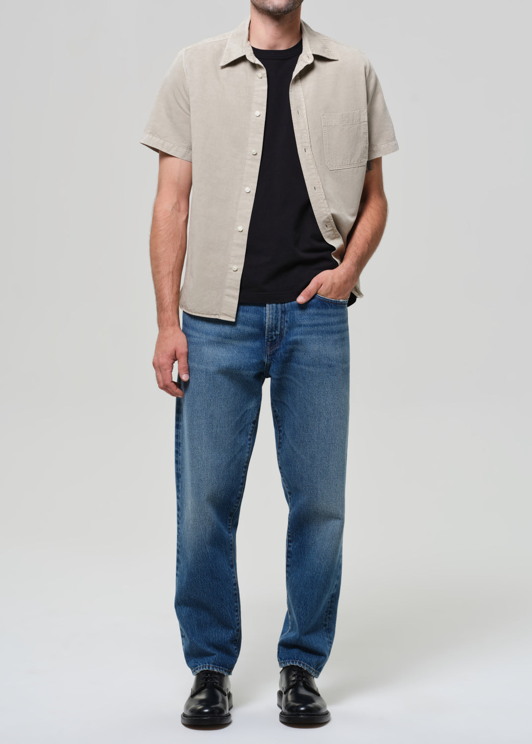 Man wearing a beige short-sleeve shirt over a black t-shirt, blue jeans, and black shoes on a grey background