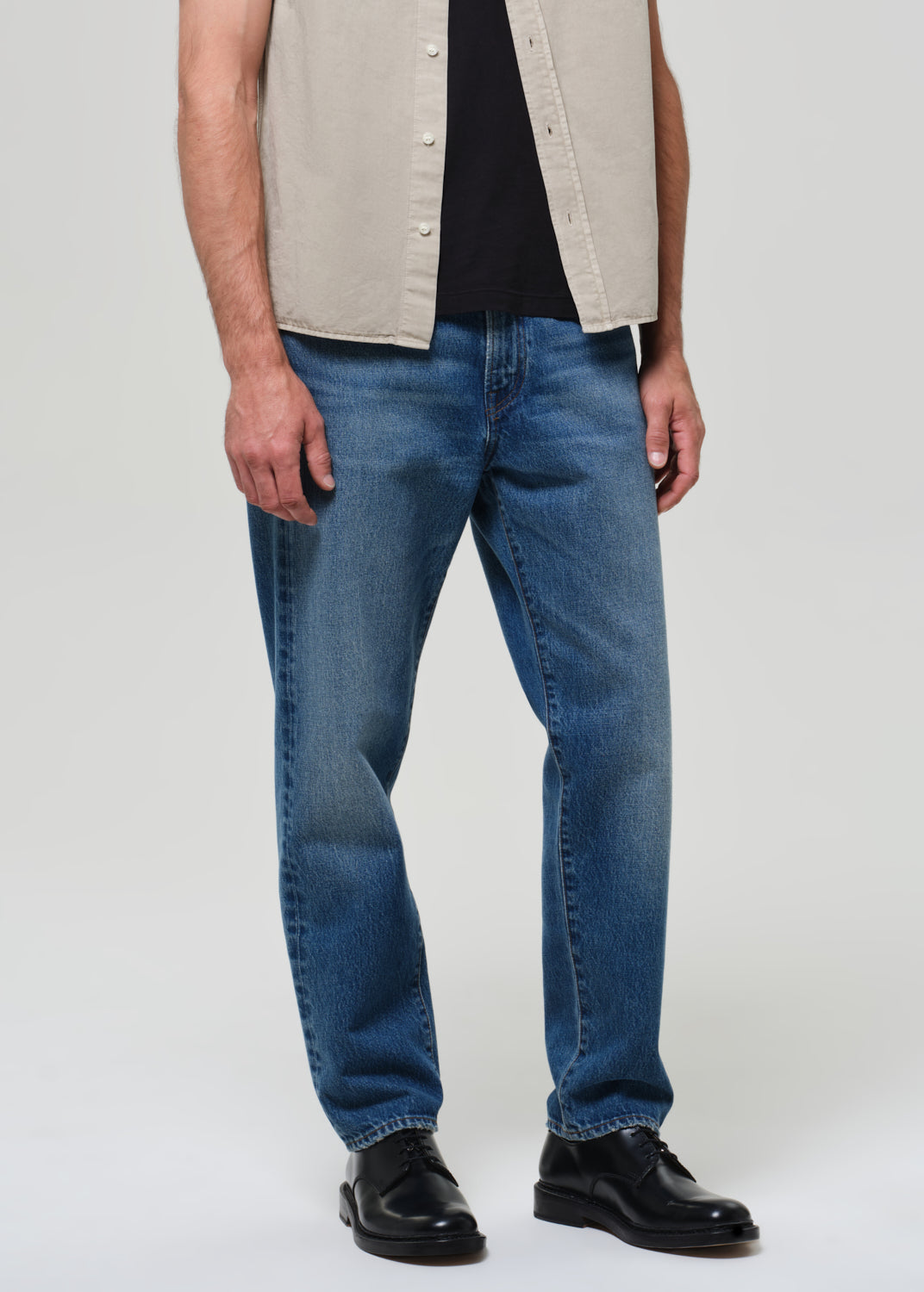 Man wearing a beige short-sleeve shirt over a black t-shirt, blue jeans, and black shoes on a grey background