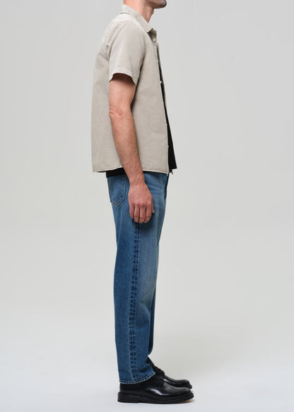 Man wearing a beige short-sleeve shirt over a black t-shirt, blue jeans, and black shoes on a grey background