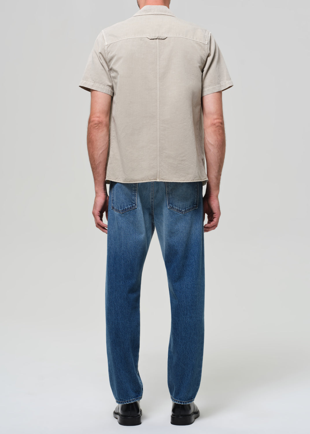 Man wearing a beige short-sleeve shirt over a black t-shirt, blue jeans, and black shoes on a grey background