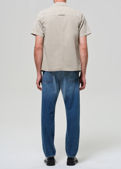 Man wearing a beige short-sleeve shirt over a black t-shirt, blue jeans, and black shoes on a grey background