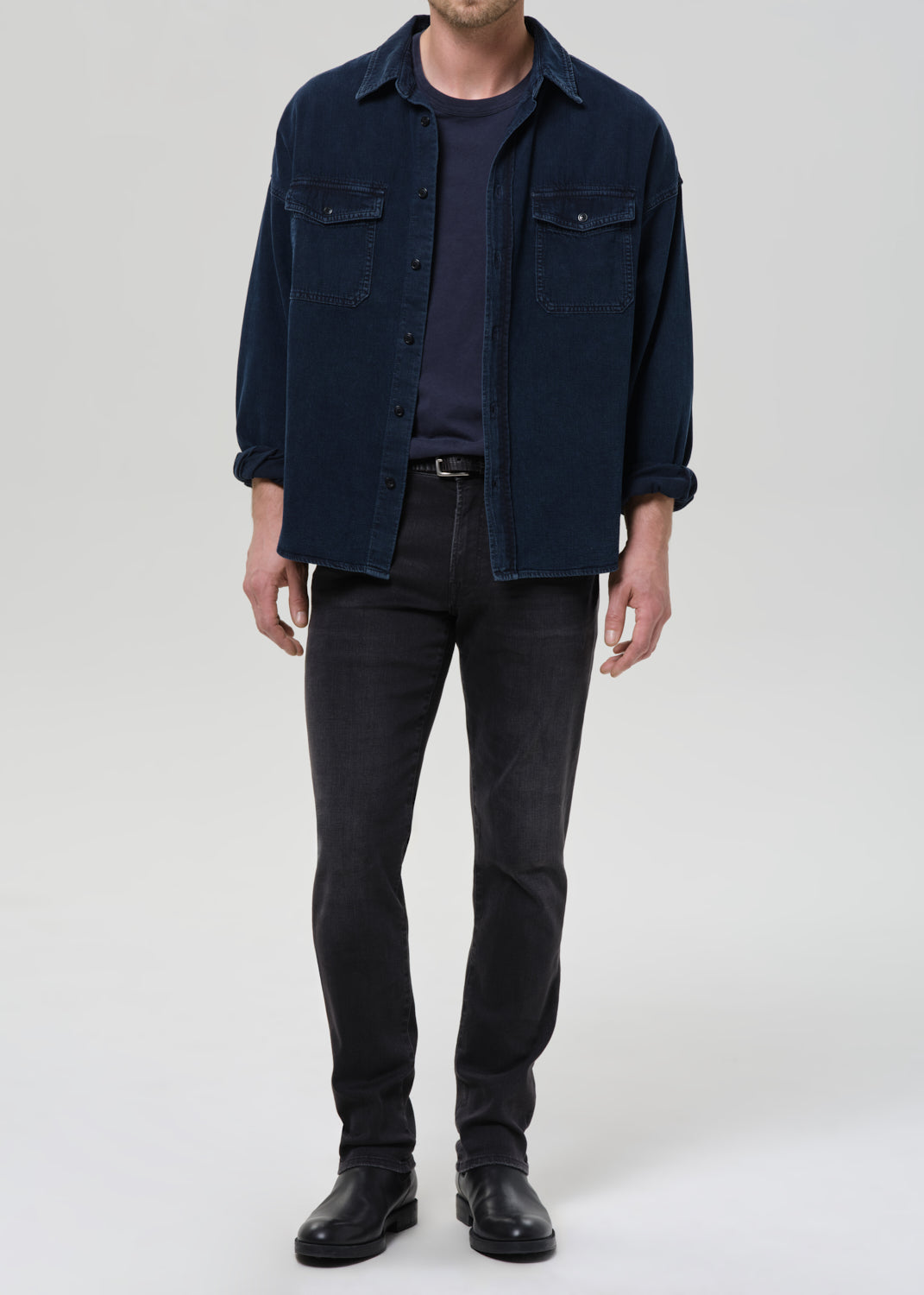 A man wearing a dark navy blue corduroy shirt jacket over a dark navy t-shirt, paired with dark grey slim-fit jeans and black leather boots, standing against a grey background.