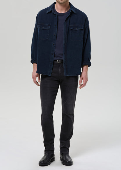 A man wearing a dark navy blue corduroy shirt jacket over a dark navy t-shirt, paired with dark grey slim-fit jeans and black leather boots, standing against a grey background.