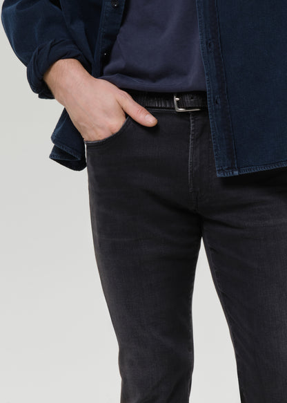 A close-up of a person wearing slim-fit dark charcoal grey jeans, a black belt, a navy t-shirt, and a dark blue overshirt, with one hand in the pocket, against a grey background.
