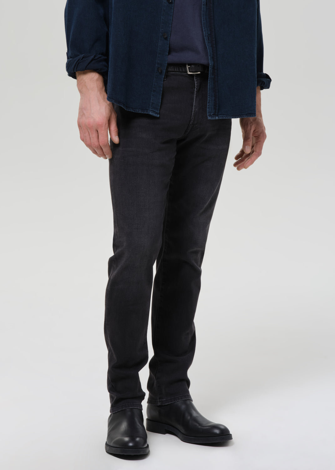A person wearing slim-fit dark charcoal grey jeans, a navy blue button-up shirt, and black leather boots, shown from the waist down against a grey background.