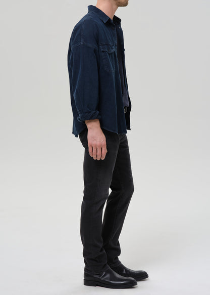 A man shown in profile wearing a dark navy corduroy button-up shirt, black trousers, and black leather shoes against a grey background.