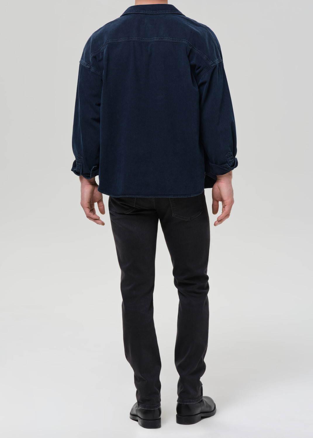 A man viewed from behind wearing a dark navy blue corduroy shirt jacket and black jeans, standing against a grey background.