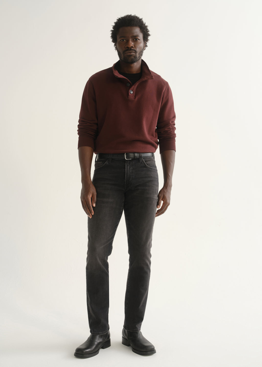 Person wearing a burgundy long sleeve polo shirt, washed black jeans, and black boots, front full body view on a light grey background.