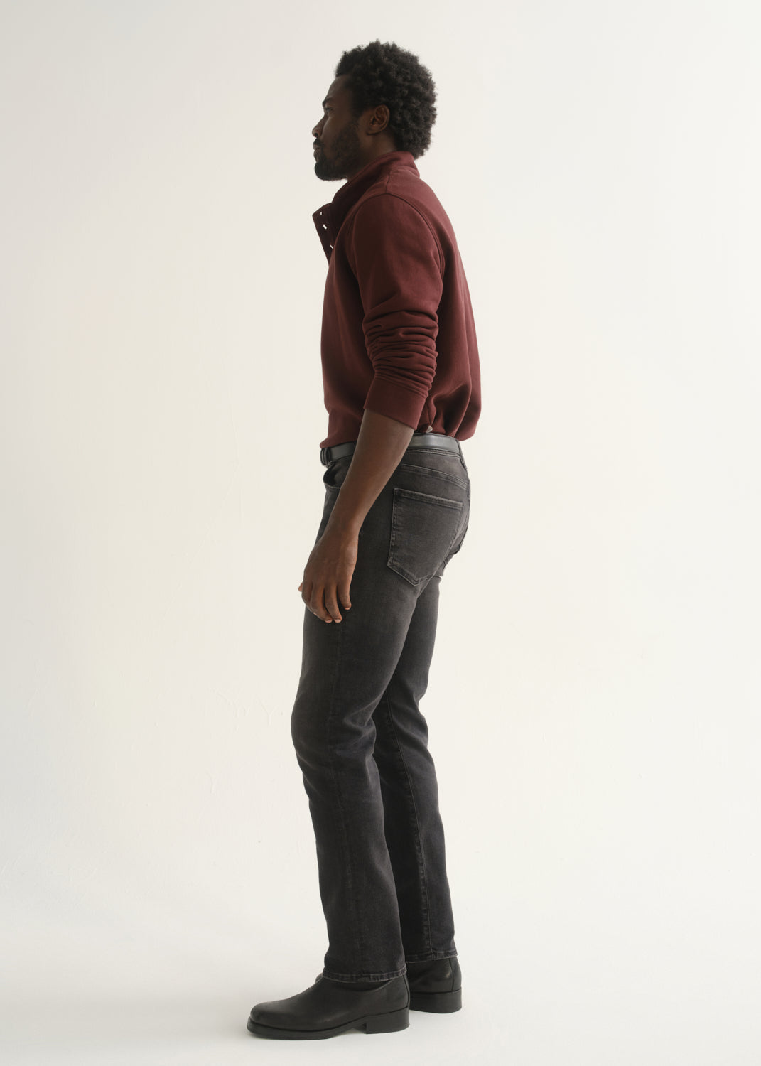 Person wearing a burgundy long sleeve polo shirt, washed black jeans, and black boots, side full body view on a light grey background.