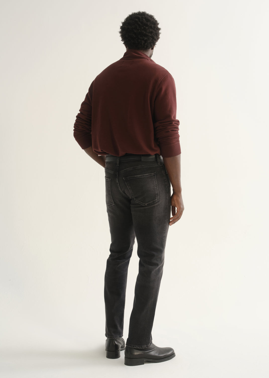 Person wearing a burgundy long sleeve polo shirt, washed black jeans, and black boots, back full body view on a light grey background.