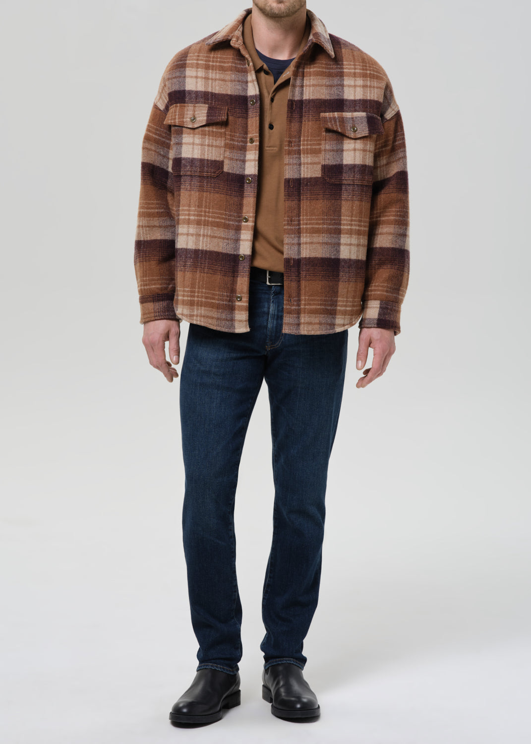 A man wearing a brown and tan plaid overshirt over a brown shirt, dark blue jeans, and black leather boots, standing against a grey background.