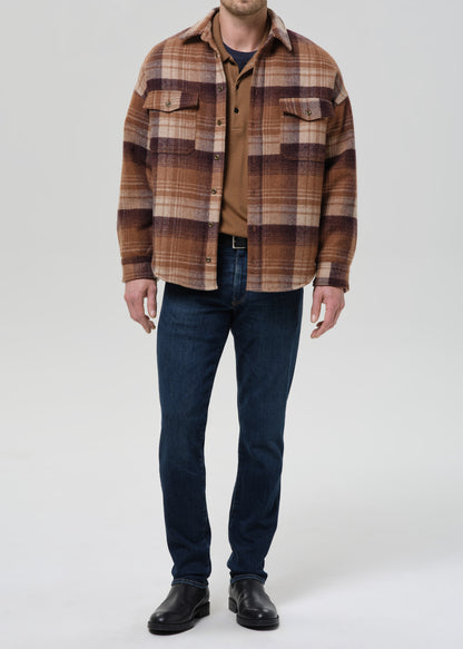 A man wearing a brown and tan plaid overshirt over a brown shirt, dark blue jeans, and black leather boots, standing against a grey background.