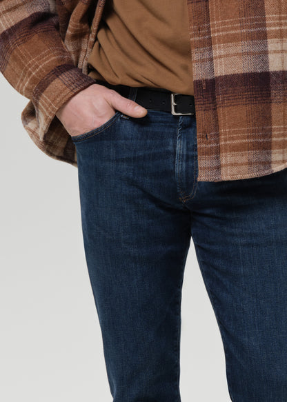 A close-up of a person wearing dark blue jeans, a black belt, a brown t-shirt, and a brown and beige plaid flannel shirt, with one hand in the pocket.