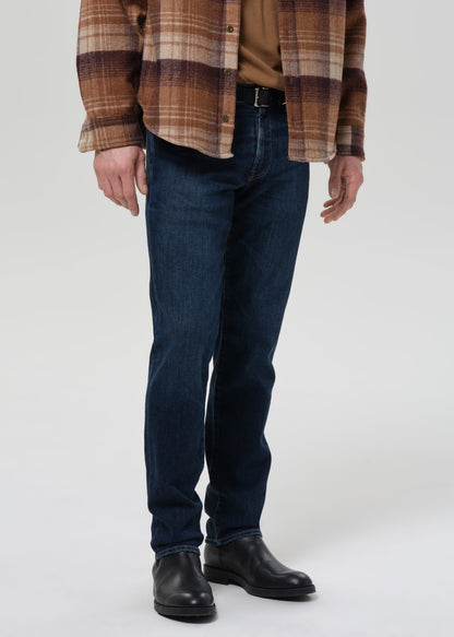 A person wearing dark blue slim-fit jeans, a brown and beige plaid flannel shirt, a tan undershirt, and black leather boots, shown from the chest down against a grey background.