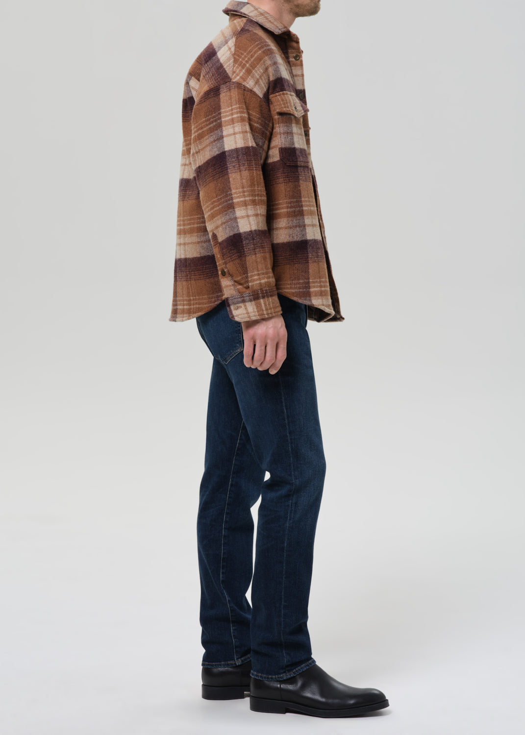 A man shown in profile wearing a brown and beige plaid flannel shirt, dark blue jeans, and black leather boots, standing against a grey background.