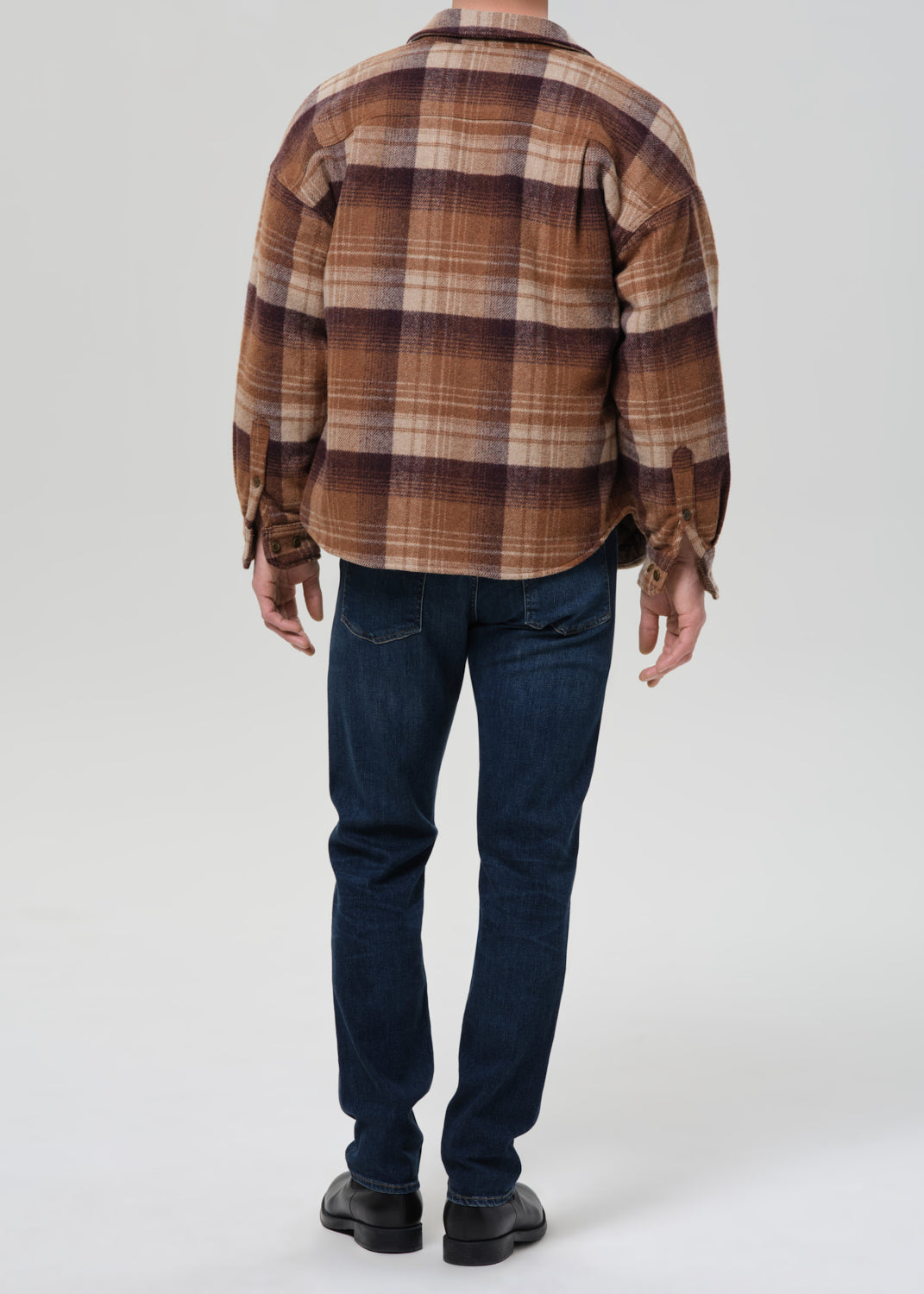 A man viewed from behind wearing a brown and beige plaid flannel shirt, dark blue jeans, and black shoes, standing against a grey background.