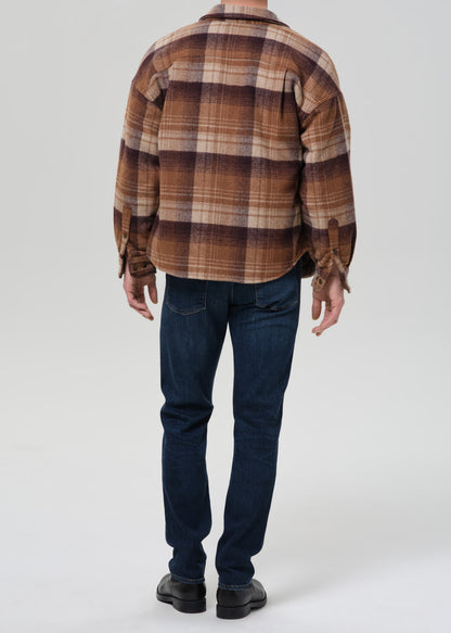 A man viewed from behind wearing a brown and beige plaid flannel shirt, dark blue jeans, and black shoes, standing against a grey background.