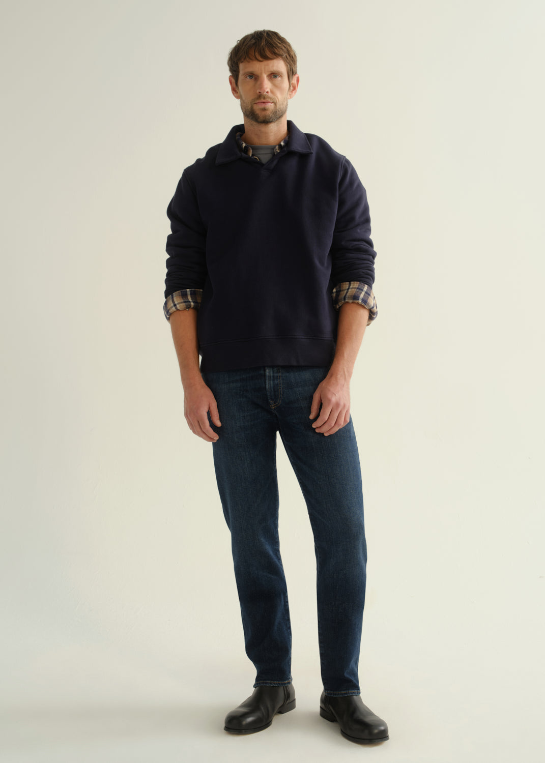 Person wearing a navy collared pullover sweatshirt layered over a plaid shirt, dark blue slim straight jeans, and black leather boots, front full body view on a light grey background