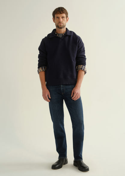 Person wearing a navy collared pullover sweatshirt layered over a plaid shirt, dark blue slim straight jeans, and black leather boots, front full body view on a light grey background