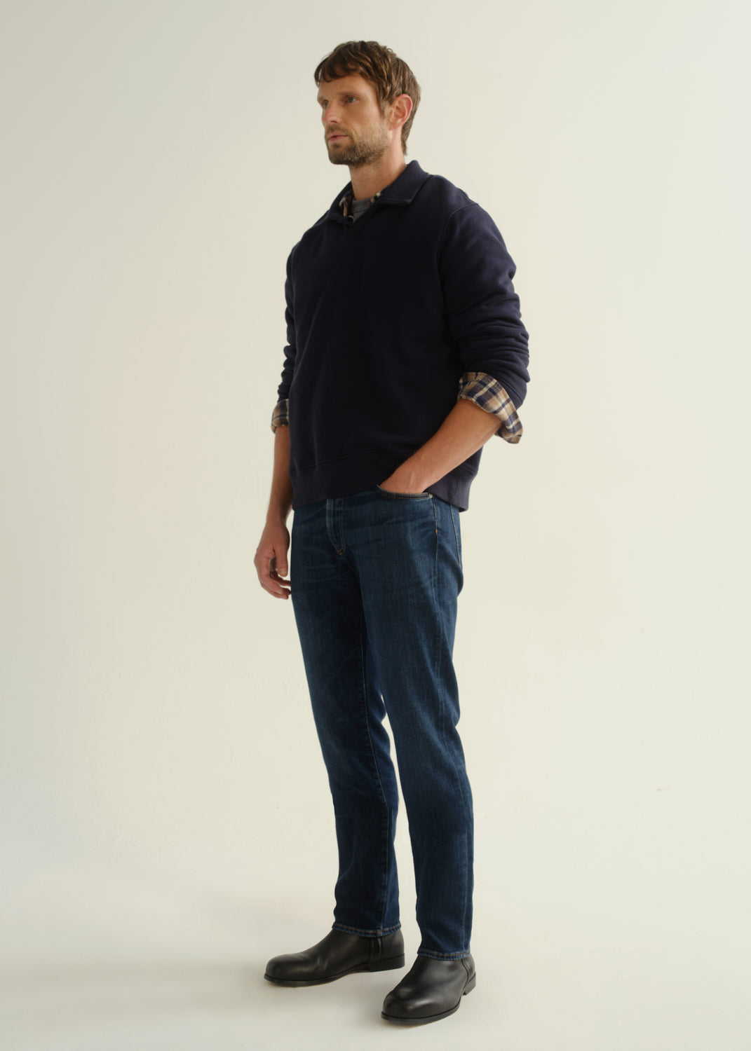 Person wearing a navy collared pullover sweatshirt layered over a plaid shirt, dark blue slim straight jeans, and black leather boots, angled front full body view on a light grey background.