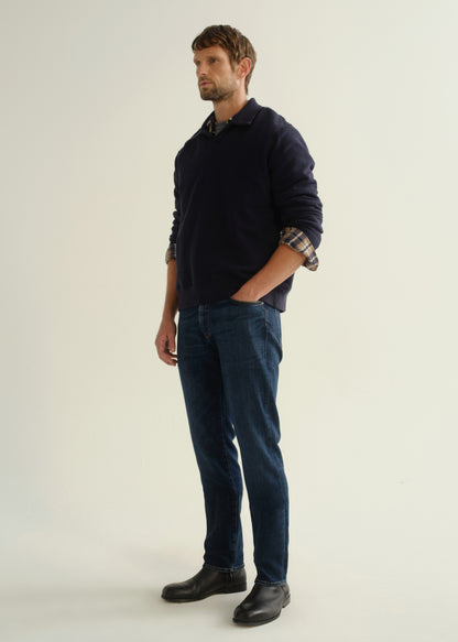 Person wearing a navy collared pullover sweatshirt layered over a plaid shirt, dark blue slim straight jeans, and black leather boots, angled front full body view on a light grey background.