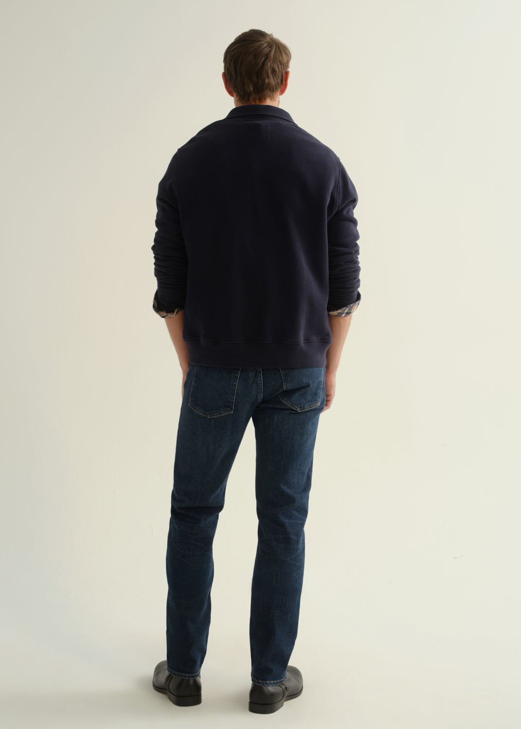 Person wearing a navy collared pullover sweatshirt layered over a plaid shirt, dark blue slim straight jeans, and black leather boots, back full body view on a light grey background.