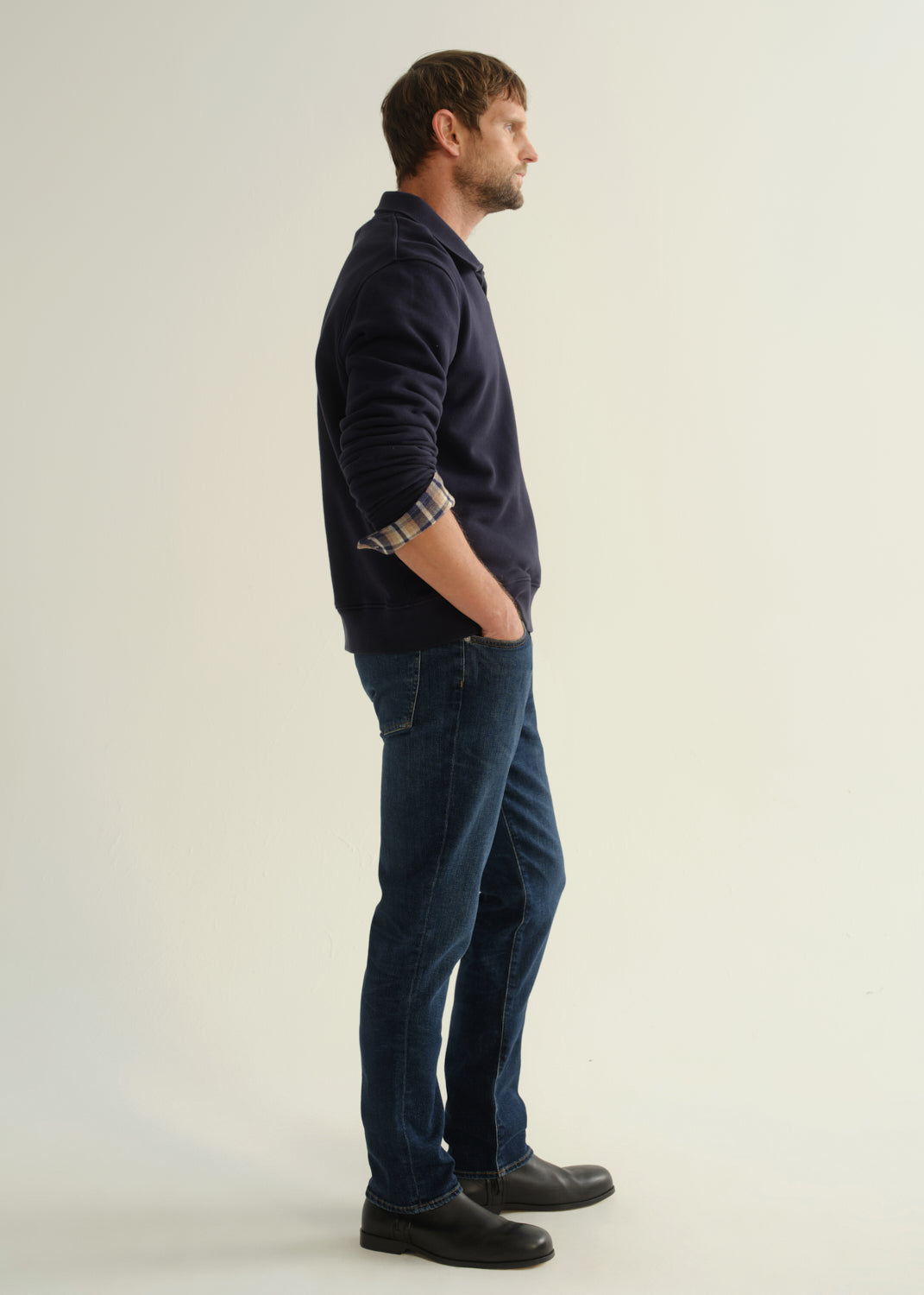 Person wearing a navy collared pullover sweatshirt layered over a plaid shirt, dark blue slim straight jeans, and black leather boots, side full body view on a light grey background.