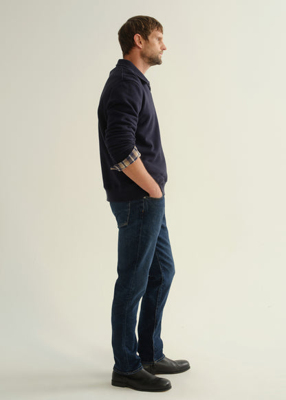 Person wearing a navy collared pullover sweatshirt layered over a plaid shirt, dark blue slim straight jeans, and black leather boots, side full body view on a light grey background.