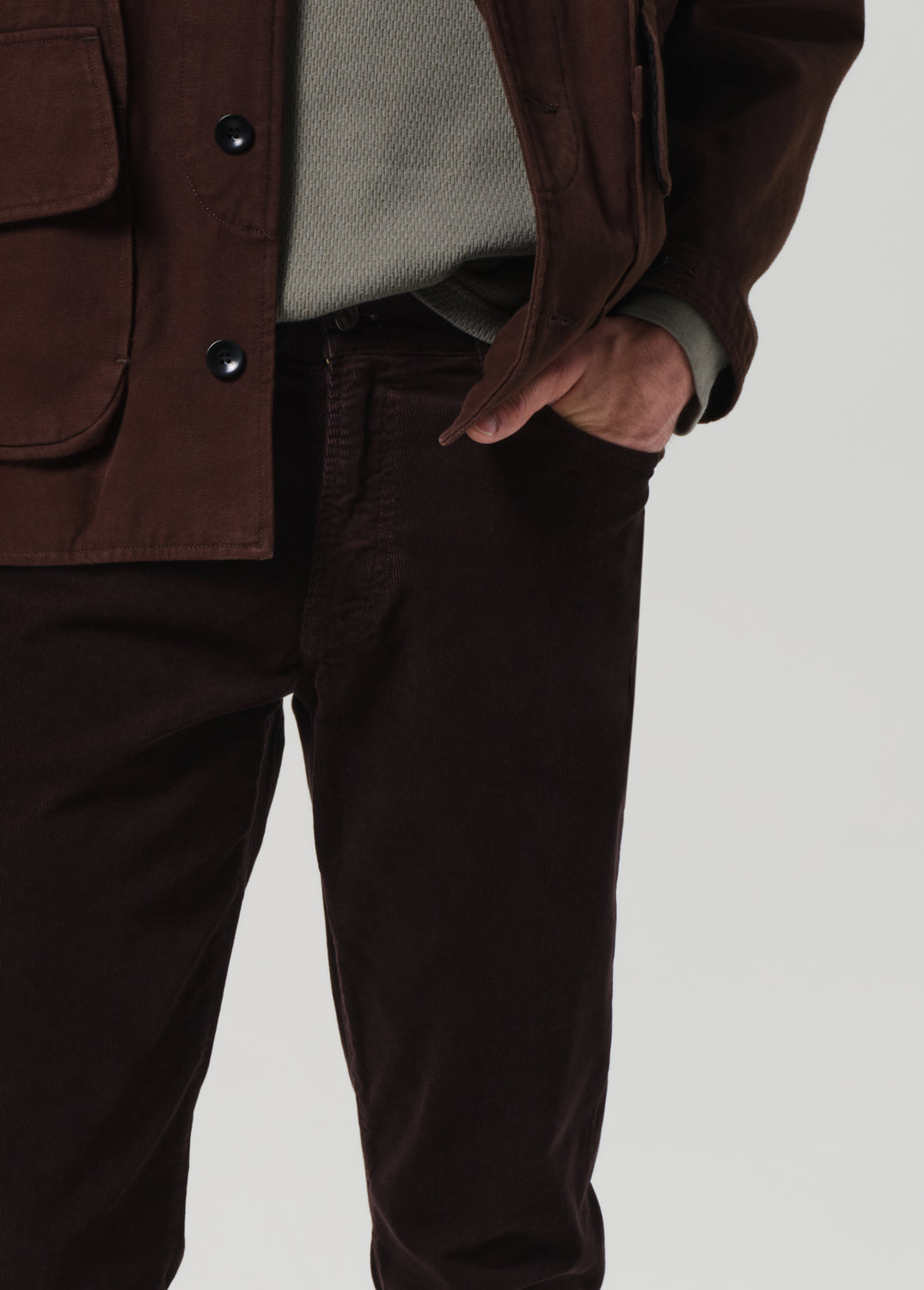 Person wearing dark brown jacket and pants with hands in pockets, gray shirt visible.