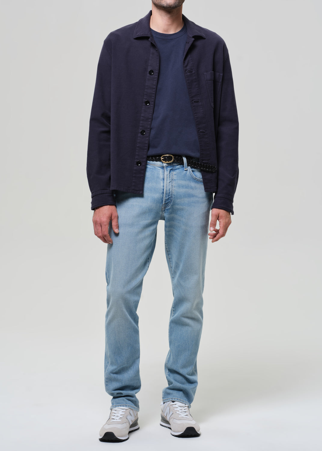 Person wearing a navy jacket, dark shirt, light blue jeans, and white sneakers on a plain background