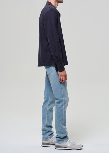 Person wearing a navy jacket, dark shirt, light blue jeans, and sneakers on a plain background