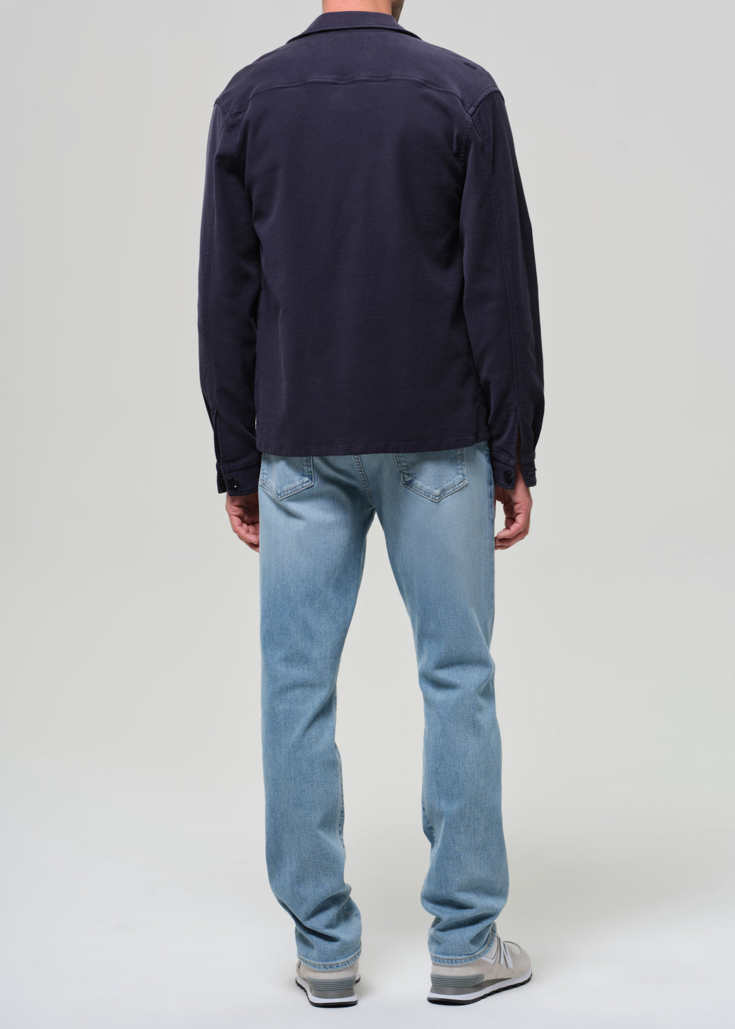 Person wearing a navy jacket, dark shirt, light blue jeans, and white sneakers on a plain background