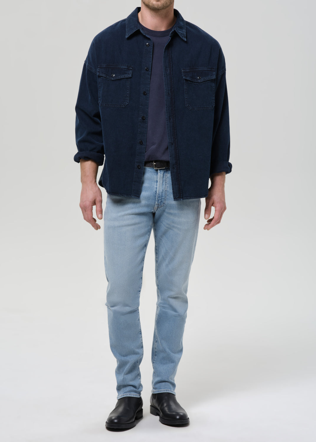 A man wearing a dark navy blue button-up overshirt over a dark t-shirt, paired with light blue jeans and black leather shoes, standing against a grey background.