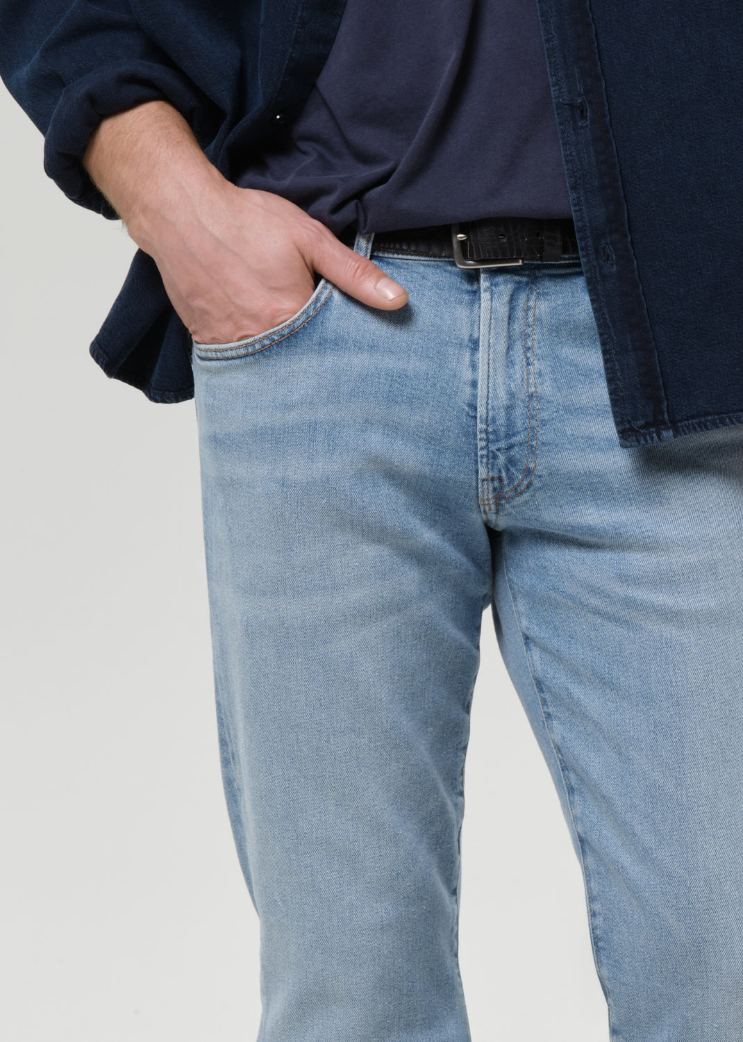 A close-up of a person wearing light blue denim jeans with a black belt, a dark navy shirt, and a dark overshirt, standing against a grey background.