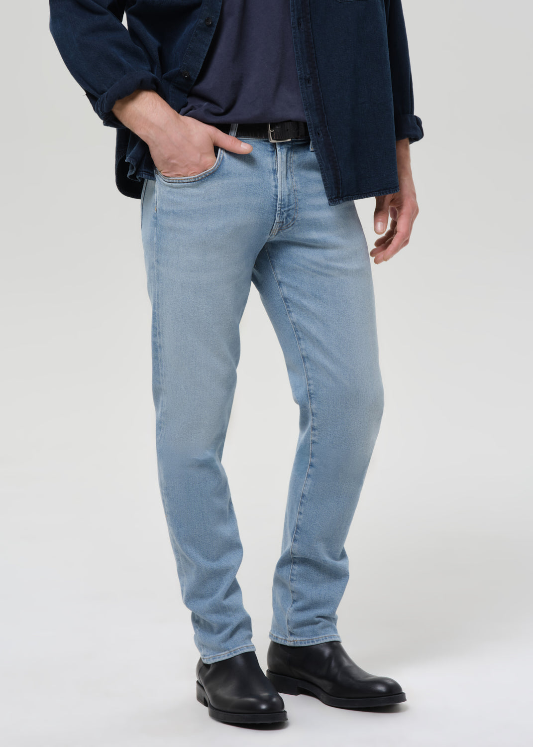 A person wearing light-wash blue slim-fit jeans, styled with a navy shirt, dark overshirt, black belt, and black leather boots, photographed against a grey background.