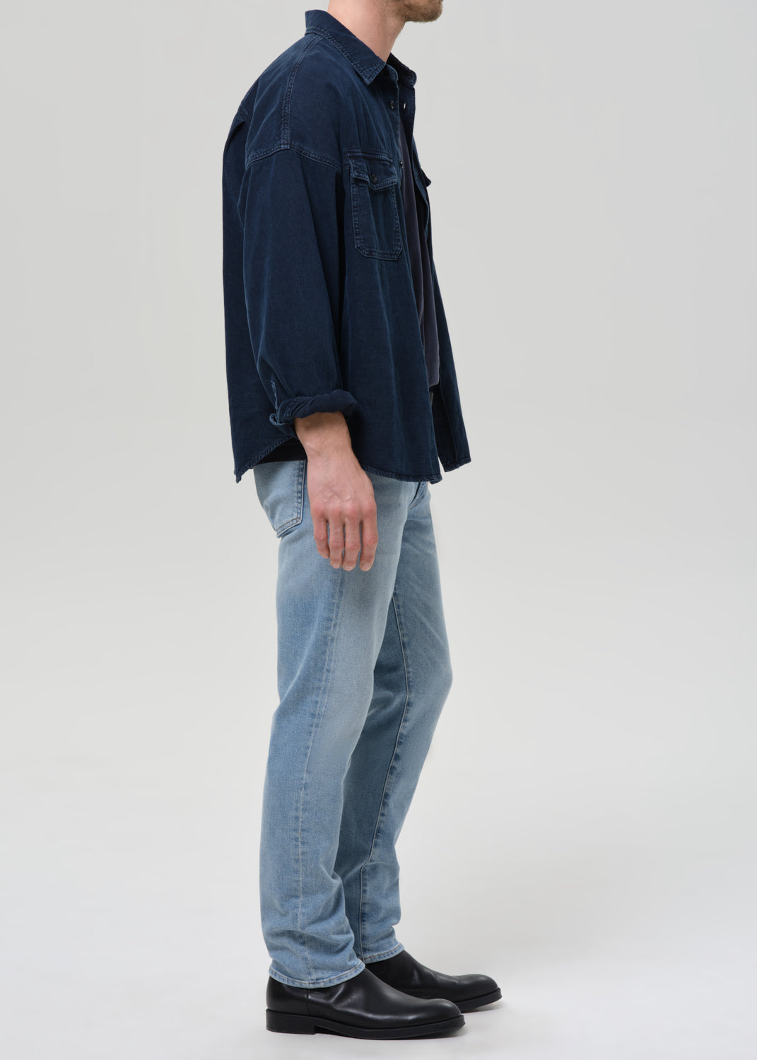 A man shown in profile wearing a dark navy blue corduroy shirt jacket, light blue jeans, and black leather shoes, standing against a grey background.