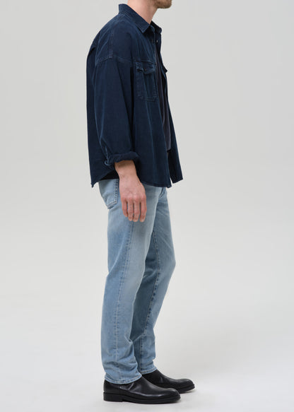 A man shown in profile wearing a dark navy blue corduroy shirt jacket, light blue jeans, and black leather shoes, standing against a grey background.