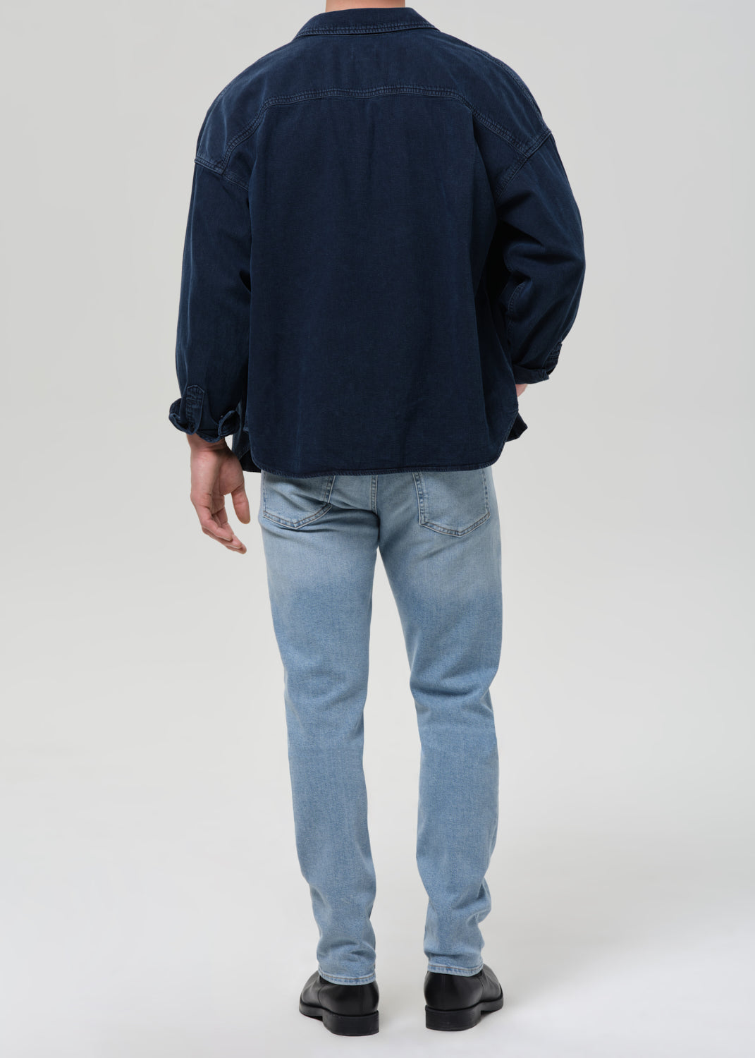 A person shown from the back wearing a dark navy overshirt, light blue jeans, and black shoes against a grey background.