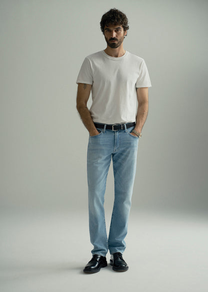 A man modeling a white crew-neck t-shirt, light-wash blue jeans, a black belt, and black leather shoes against a neutral gray studio background.