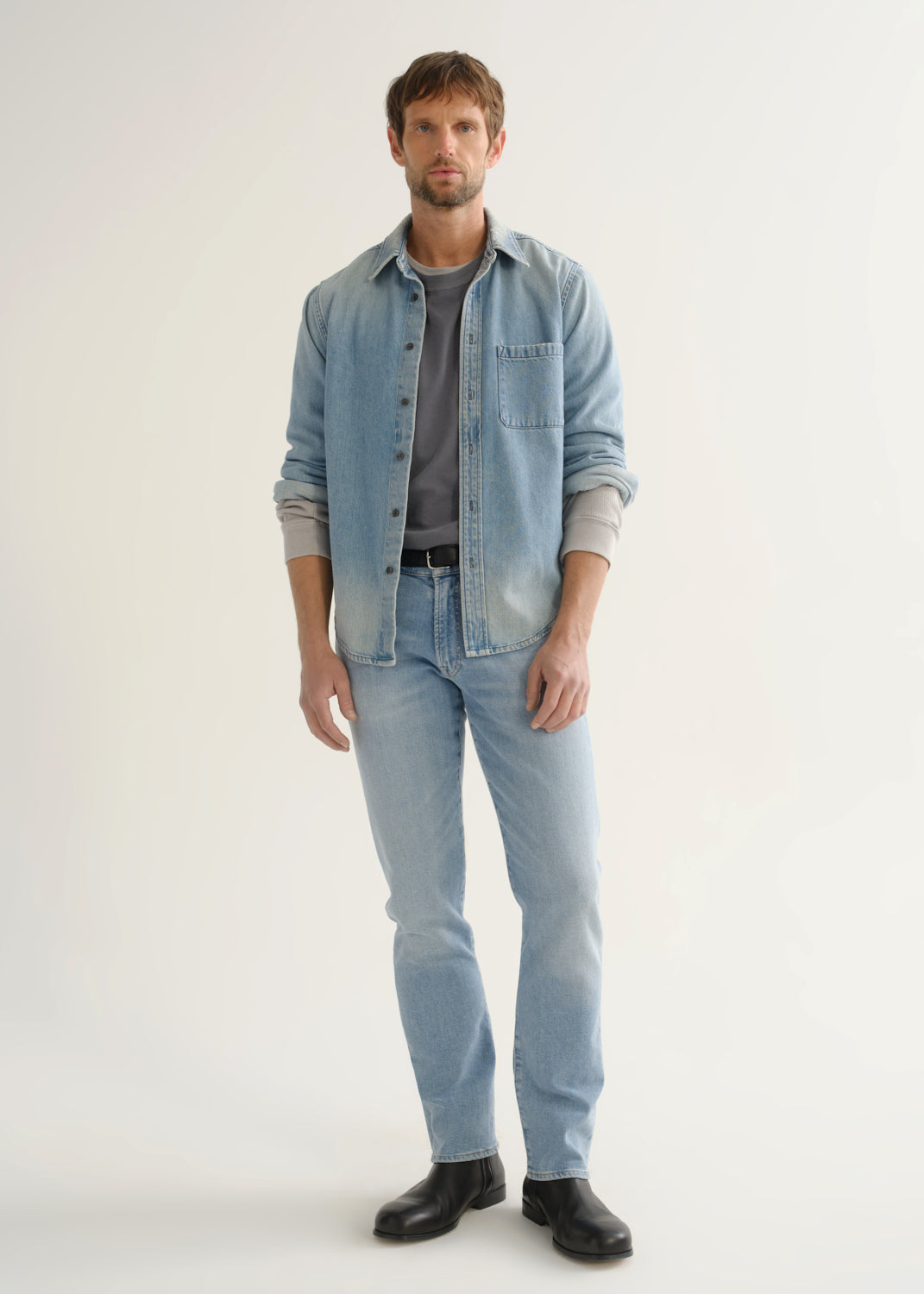 Person wearing a light blue denim shirt over a grey top, light wash blue jeans, and black shoes, front full body view on a light grey background.