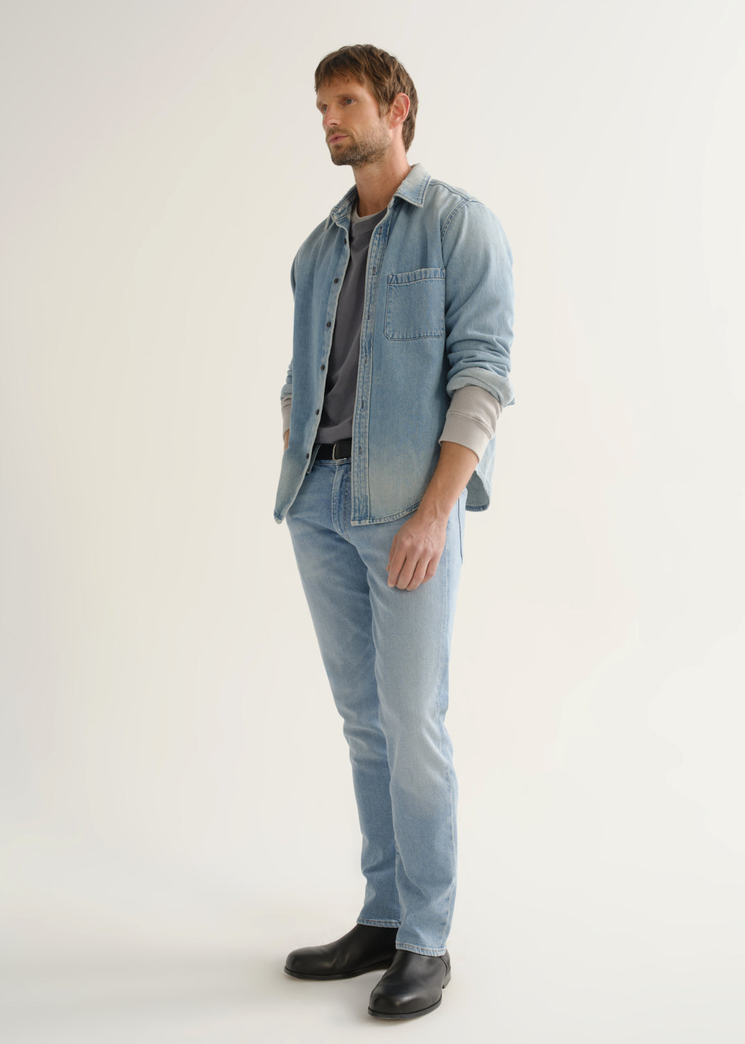 Person wearing a light blue denim shirt, light wash blue jeans, and black shoes, side full body view on a light grey background.