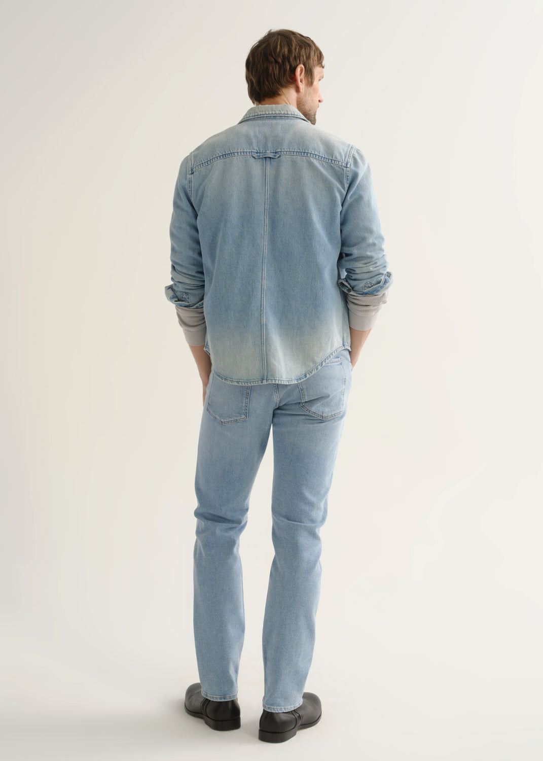 Person wearing a light blue denim shirt, light wash blue jeans, and black shoes, back full body view on a light grey background.