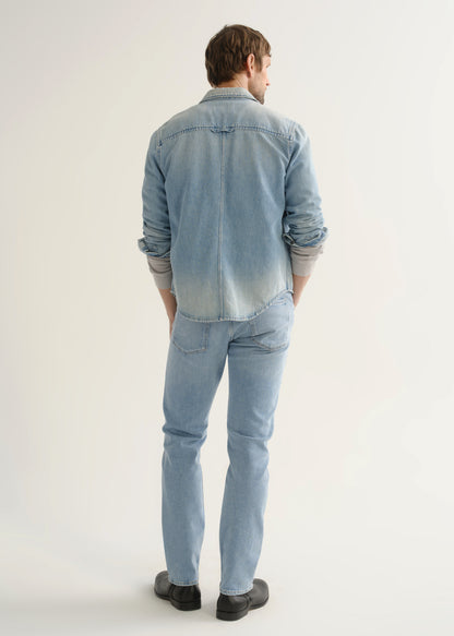 Person wearing a light blue denim shirt, light wash blue jeans, and black shoes, back full body view on a light grey background.