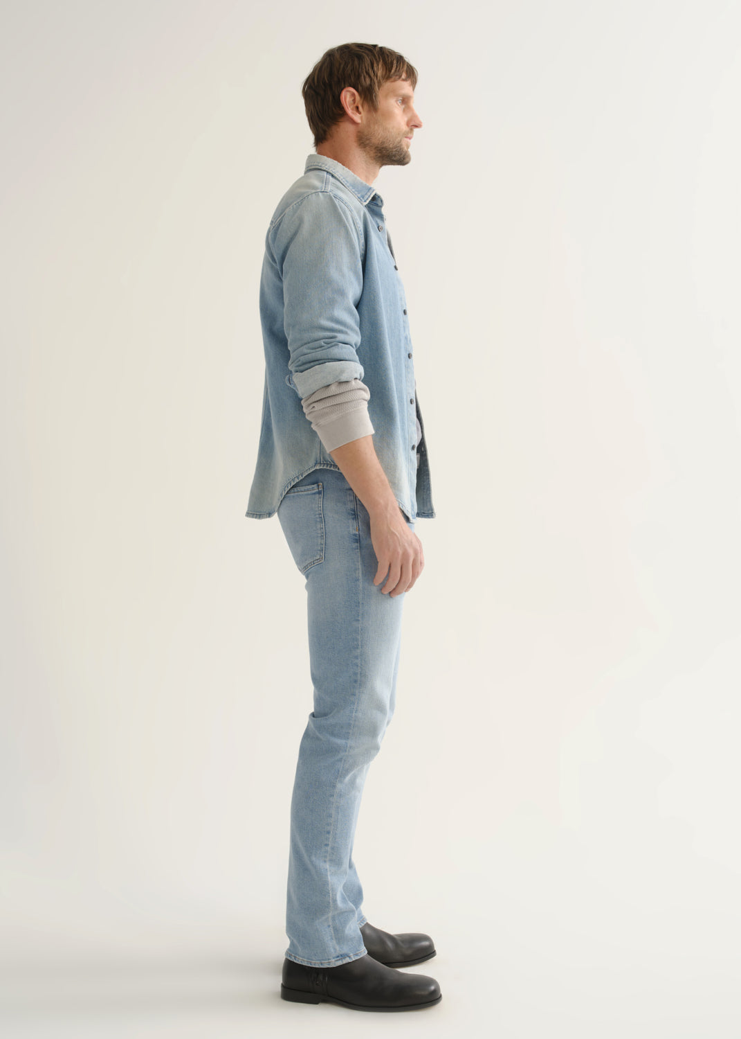 Person wearing a light blue denim shirt, light wash blue jeans, and black shoes, side full body view on a light grey background.