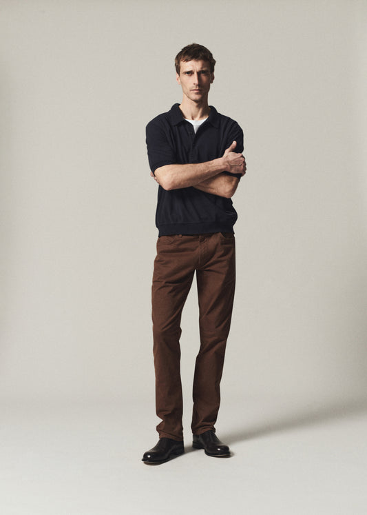 A man stands with arms crossed wearing a dark navy polo shirt, brown trousers, and black shoes against a plain light gray studio background.